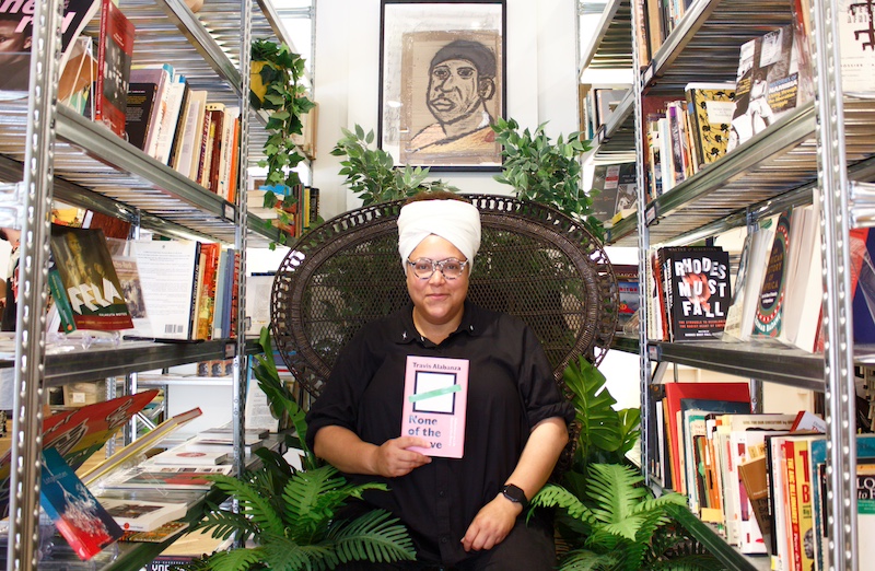 A Black non-binary person with a black shirt and jeans and a white headwrap sits in a chair in between two bookcases while holding a book titled “None of the Above”