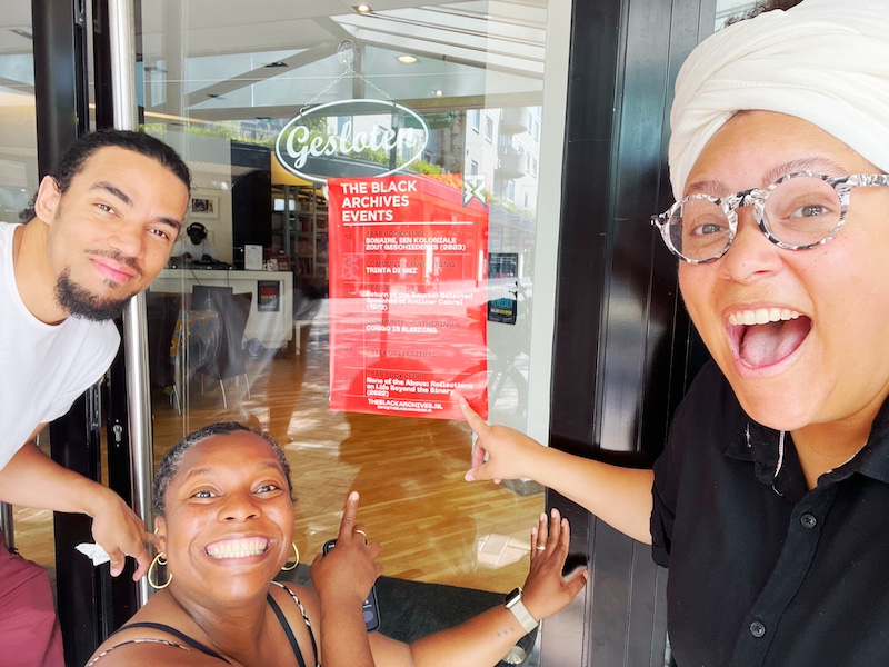 Three Black people, smile at the camera while pointing at an events poster displayed in the window of The Black Archives Bijlmer