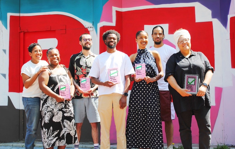 Seven people stand in front of a colorful street art wall while holding a book titled “None of the Above”