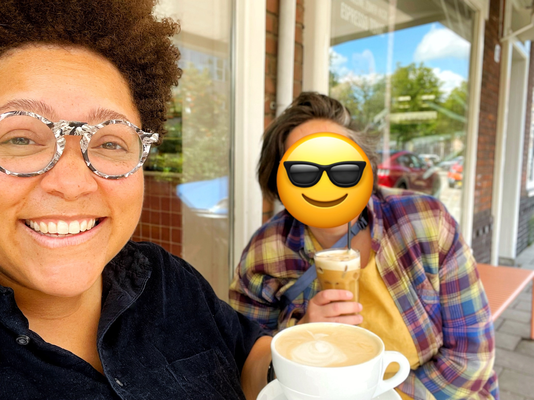 A selfie of two people smiling at the camera while holding coffee on a sunny terrace