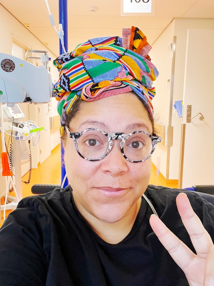 A Black person in a headwrap is hooked up to an IV drip and waves two fingers at the camera in a hospital ward