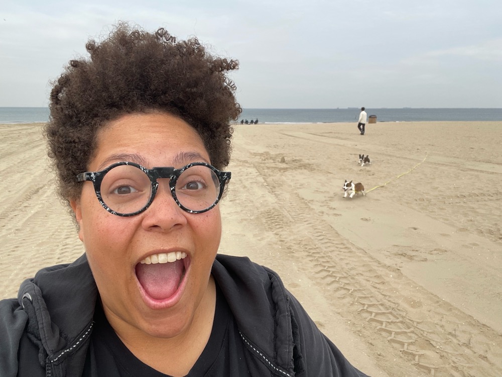 Afropean person smiling brightly at the camera while at the beach, happy dogs in the background
