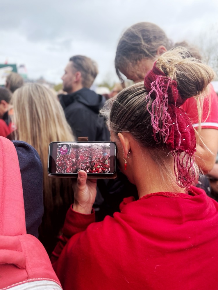 A woman listens to a live feed of the protest she’s at, the crowd being so large that the speakers can’t reach her ear