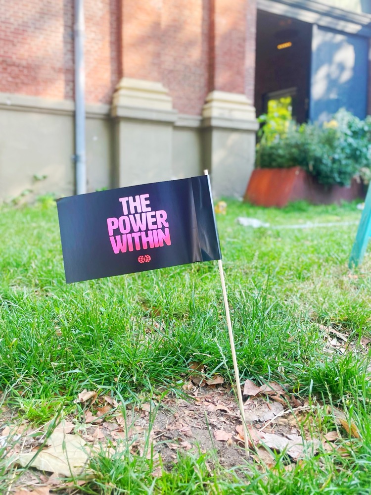 A small paper flag reading 'The Power Within' is stuck in the grass of the Amsterdam Marineterrein