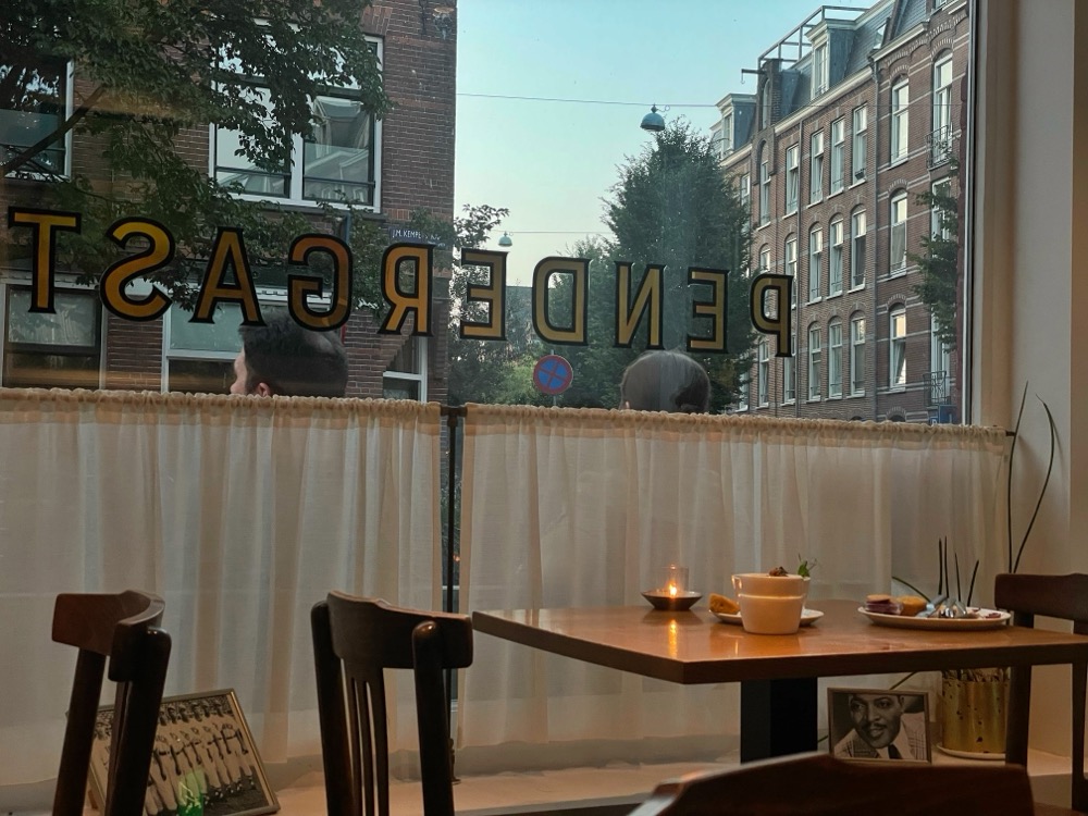 A restaurant window photographed from the inside, logo visible on the window. White curtains hang across, old-fashioned photos of Black musicians.