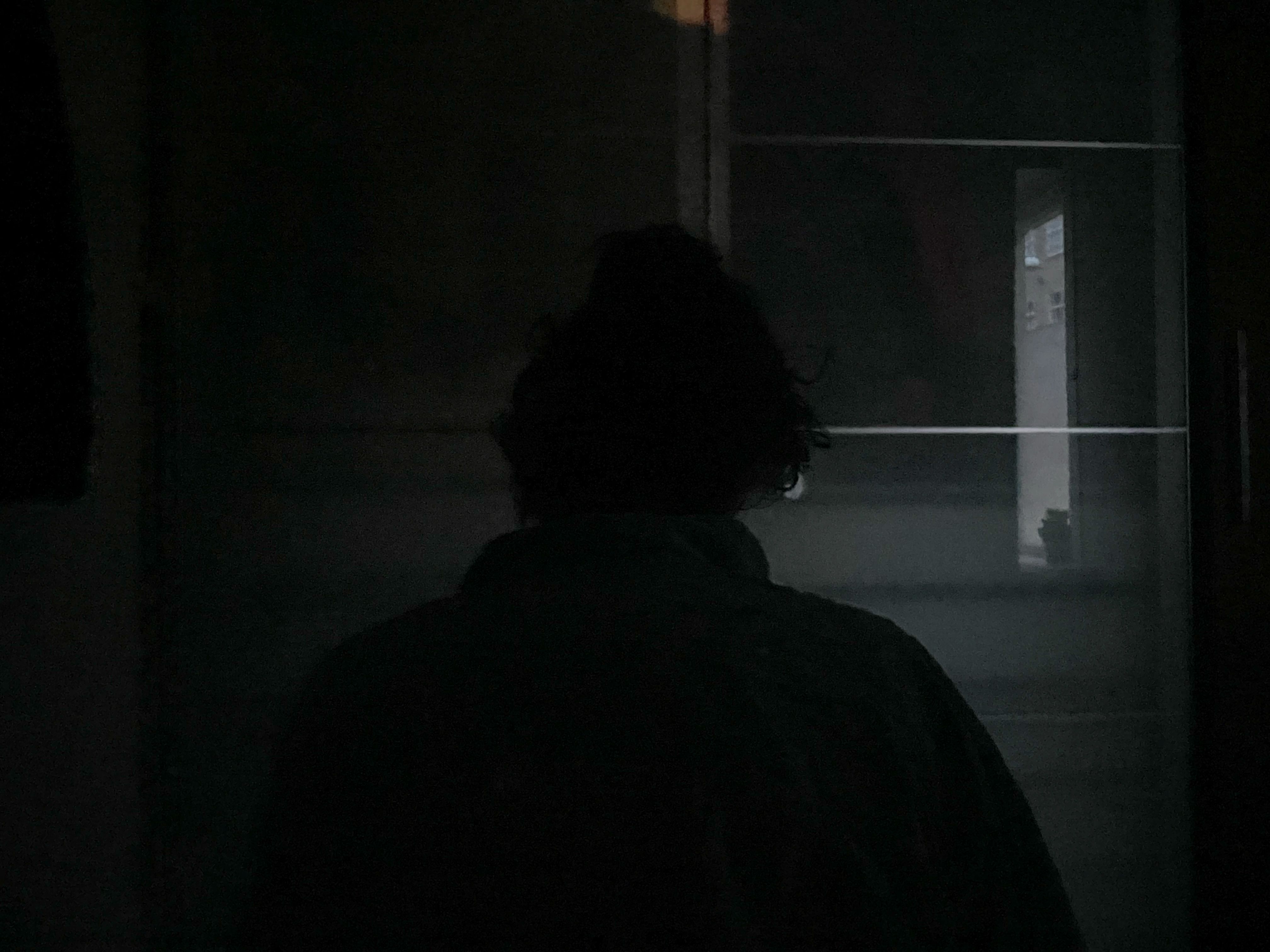 A woman sits in a dark room with her back to the camera, readying herself to get up